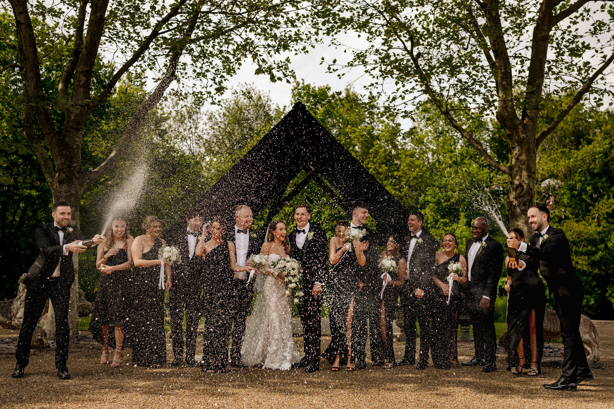 Bride and groom being sprayed with Champagne by the bridal party in black tie and green bridesmaid dresses at a Barn Wedding venue near Farnham, Bury Court Barn.